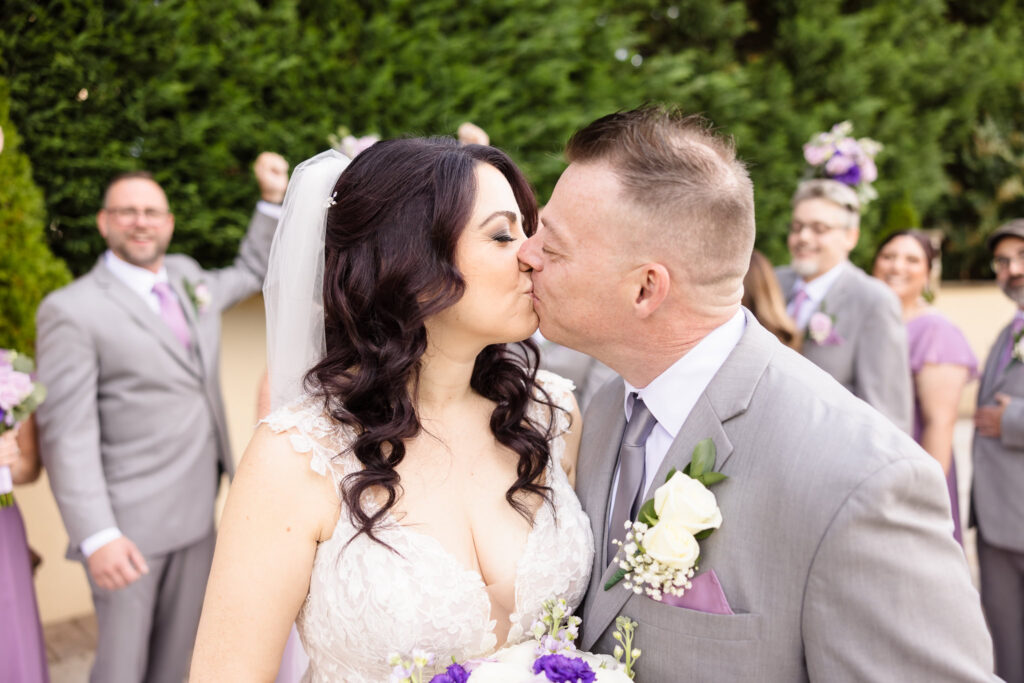 Bride and groom share a joyful kiss surrounded by cheering bridal party in lavender and gray, capturing a lively celebration moment at Larkfield.