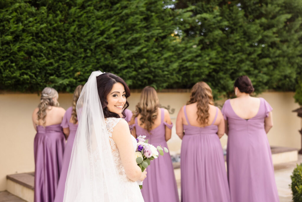 Bride smiling over her shoulder while holding a white and lavender bouquet, as her bridesmaids in matching mauve gowns await the first look moment.