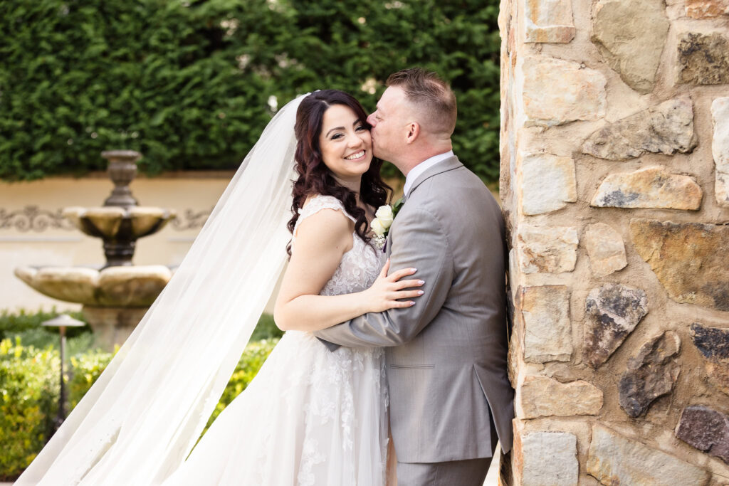Groom kissing the bride on the cheek as she smiles, standing beside a stone wall with her veil flowing behind and a garden fountain in the background.
