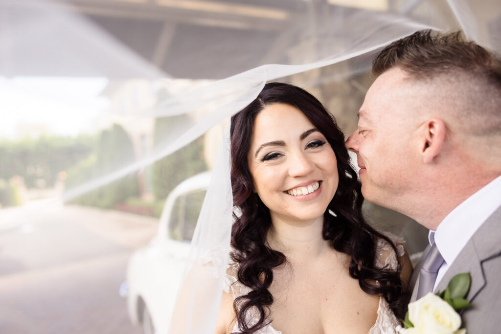 Bride and groom sharing a joyful moment under her flowing veil, with the bride smiling radiantly as a vintage white car softly blurs into the background.
