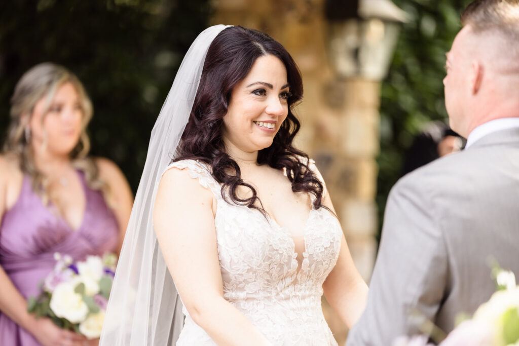 Bride smiling tenderly at the groom during their outdoor ceremony at Larkfield, with a bridesmaid in lavender holding a bouquet in the background.