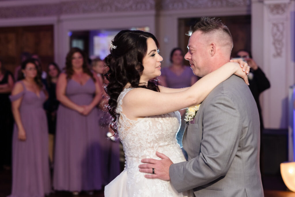 Bride and groom sharing their first dance at Larkfield, embracing under soft lighting as guests in lavender dresses look on with emotion and joy.