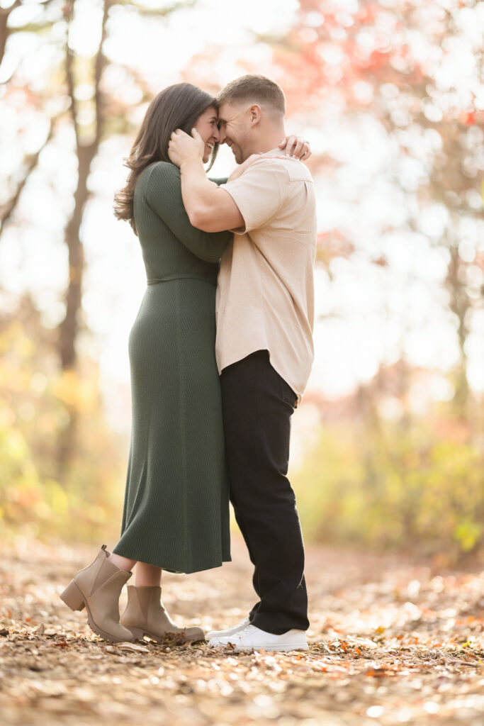 Engaged couple standing forehead to forehead on a sun-dappled trail at Bethpage State Park, surrounded by soft autumn light and fallen leaves — a quiet moment of warmth and connection.