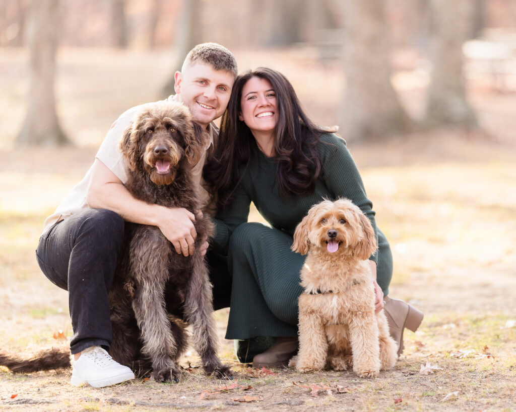 Couple kneeling with their two smiling dogs during a sunlit engagement session at Bethpage State Park, capturing a joyful and relaxed fall moment.