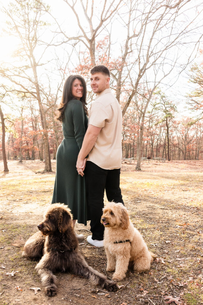 Couple holding hands and smiling at the camera while their two dogs sit contentedly at their feet, surrounded by leafless trees and golden sunlight at Bethpage State Park.