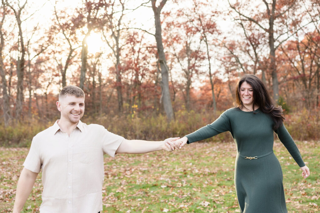 Couple laughing and holding hands as they walk through a leaf-strewn meadow at Bethpage State Park, framed by soft golden light and quiet autumn trees.