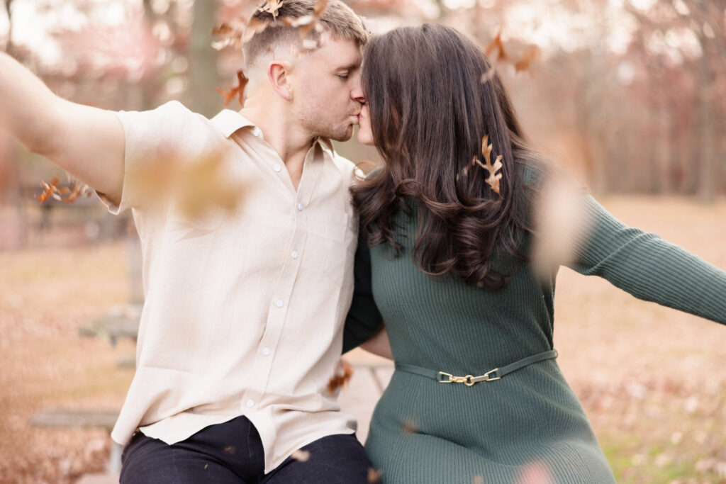 Engaged couple sharing a playful kiss as autumn leaves fall around them at Bethpage State Park capturing spontaneity, romance, and late fall charm.
