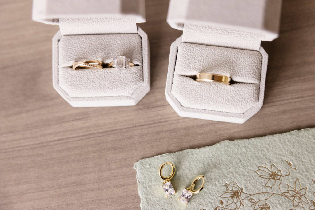 Flat lay of wedding rings and gold earrings with emerald-cut diamonds, styled in ivory velvet boxes atop a wood surface, capturing elegant bridal details.