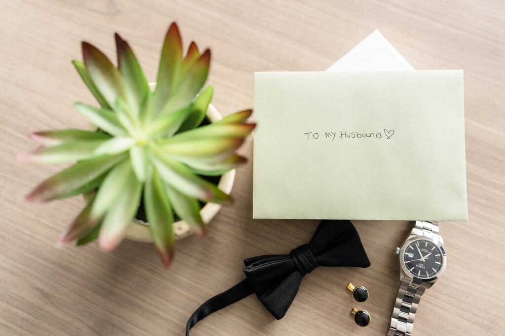 Flat lay of groom’s details featuring a handwritten “To My Husband” letter, silver wristwatch, black bow tie, cufflinks, and a potted succulent, captured during wedding morning preparations at Danfords Hotel in Port Jefferson.