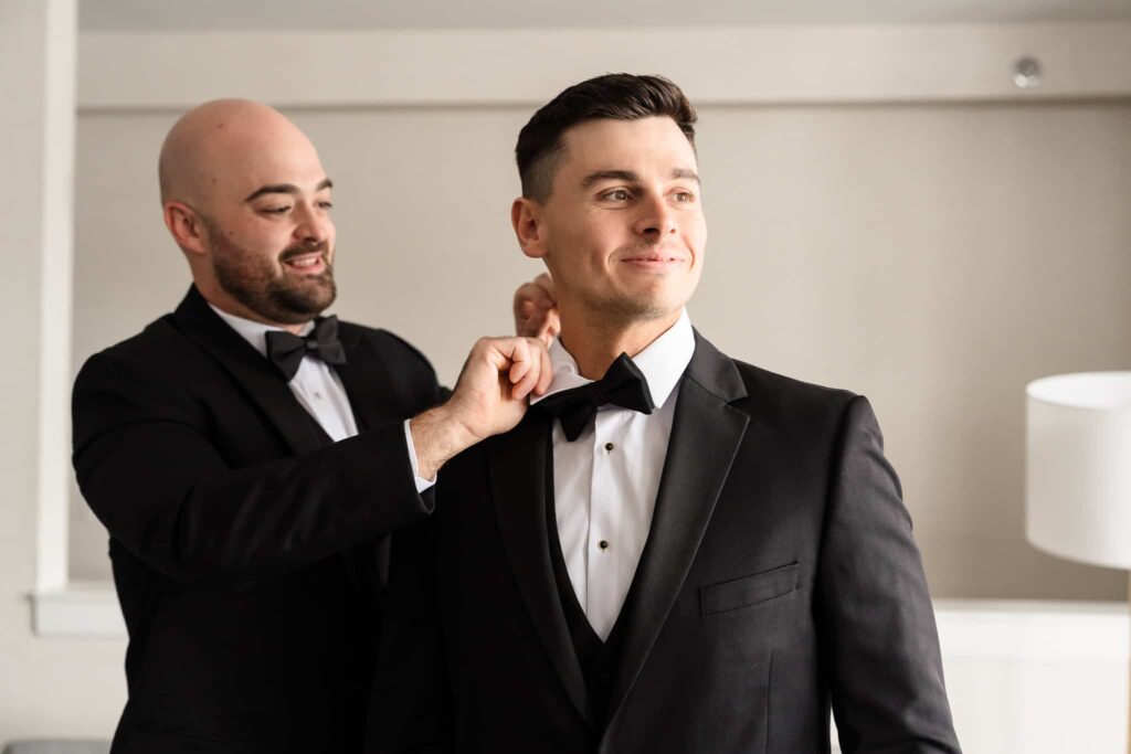 Groomsman adjusting the groom’s black bow tie as they prepare together in classic black tuxedos at Danfords Hotel before a timeless Long Island wedding at the Old Field Club.