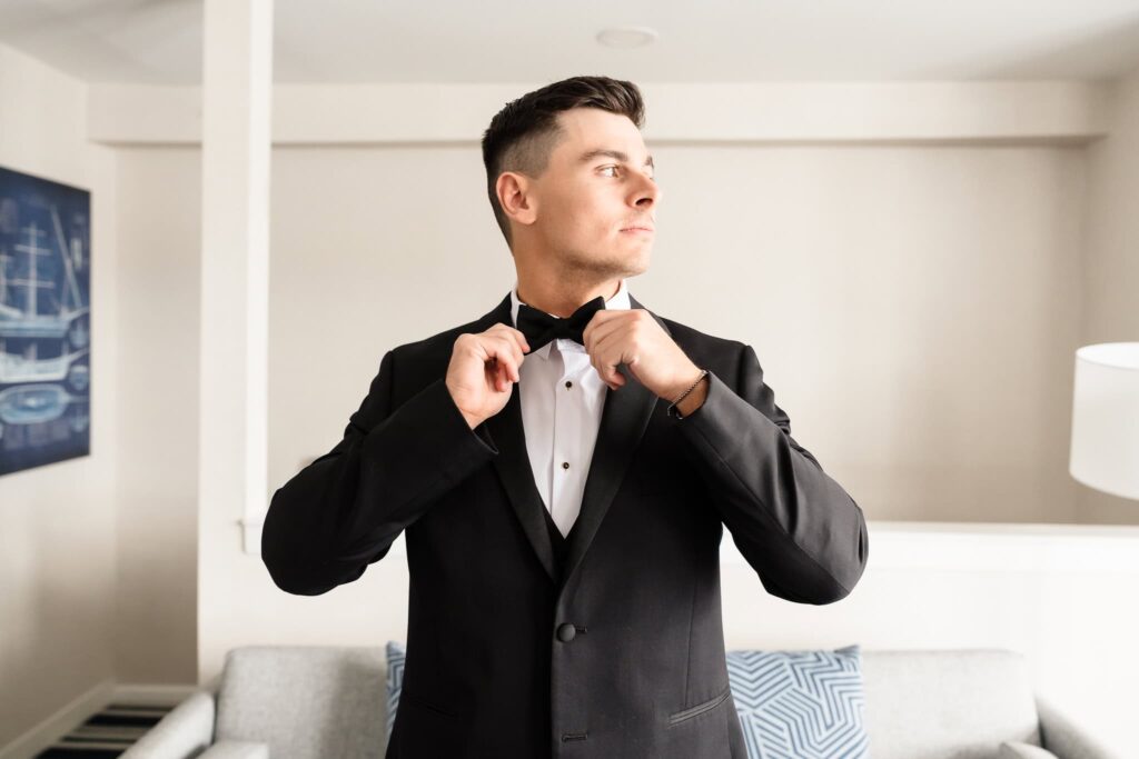 Groom adjusting his black bow tie in a light-filled suite at Danfords Hotel, dressed in a classic tuxedo as he prepares for his elegant Old Field Club wedding on Long Island.