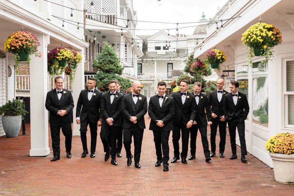 Groom walking with his groomsmen in classic black tuxedos through the charming brick courtyard of Danfords Hotel in Port Jefferson, surrounded by hanging mums and string lights before their Old Field Club wedding.