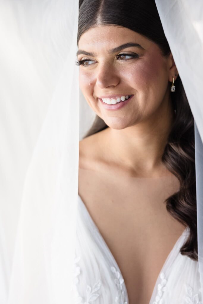 Radiant bridal portrait of the bride framed by her veil, showcasing soft makeup, gold earrings, and a delicate lace gown on the morning of her elegant Long Island wedding.
