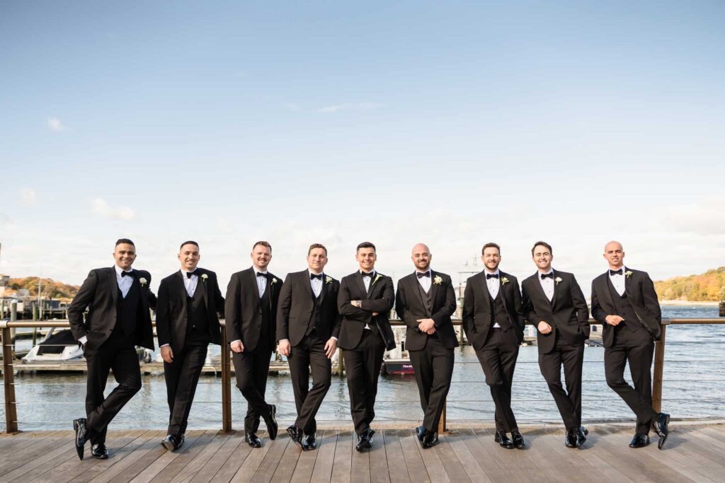 Groom and groomsmen in classic black tuxedos posing along the waterfront dock at Danfords Hotel Marina, with boats and fall foliage in the background before a North Shore Long Island wedding at the Old Field Club.