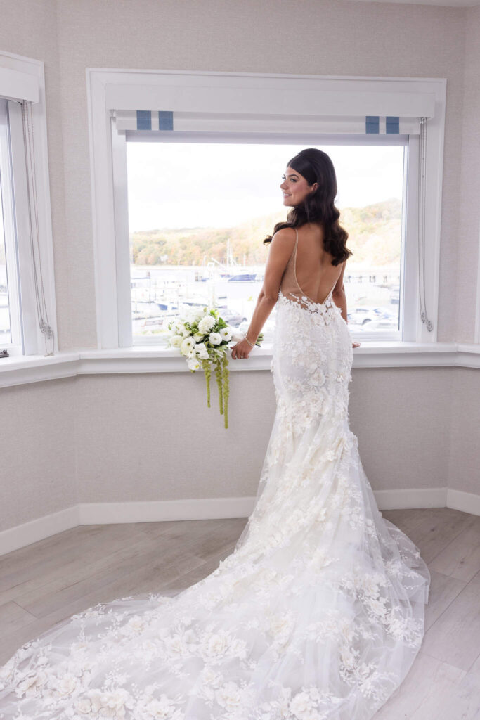 Bride standing by a window overlooking the Port Jefferson marina, holding a white bouquet and wearing a floral applique gown with a dramatic train and open back before her Old Field Club wedding on Long Island.