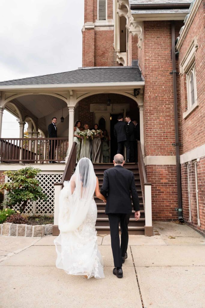 Bride walking hand-in-hand with her father up the steps of Infant Jesus Church, her veil trailing behind as bridesmaids and groomsmen wait above, moments before the ceremony begins on Long Island.