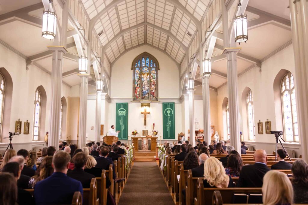 Wide view of the wedding ceremony at Infant Jesus Church in Port Jefferson, with guests seated beneath vaulted ceilings and stained glass as the priest delivers a heartfelt message at the altar.