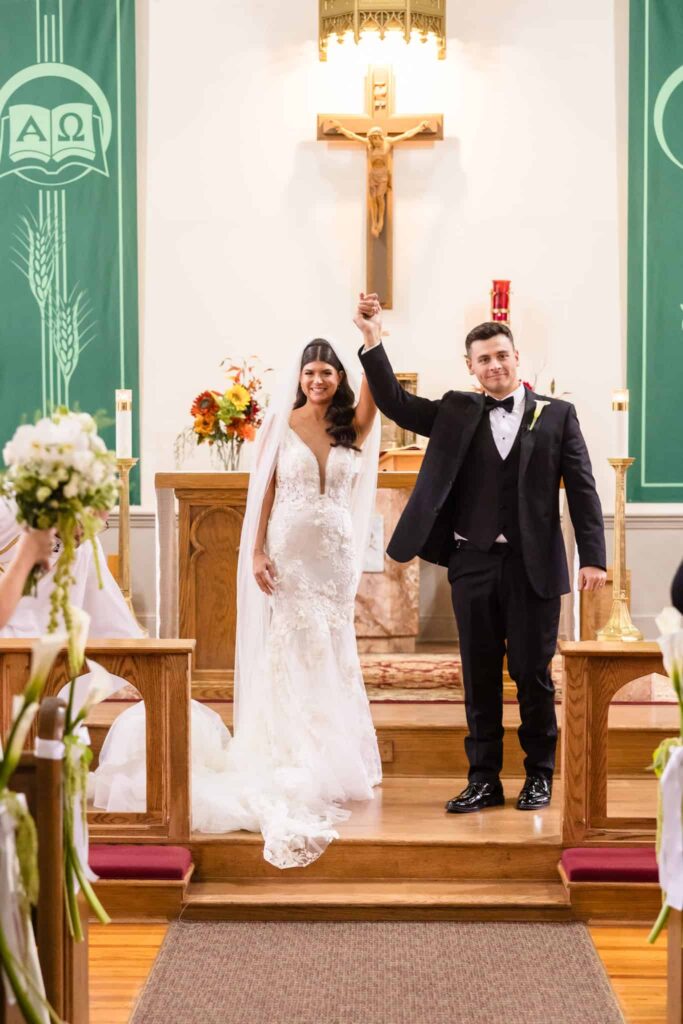 Just married bride and groom joyfully raise their joined hands at the altar of Infant Jesus Church, standing beneath the crucifix after their heartfelt Catholic wedding ceremony on Long Island.