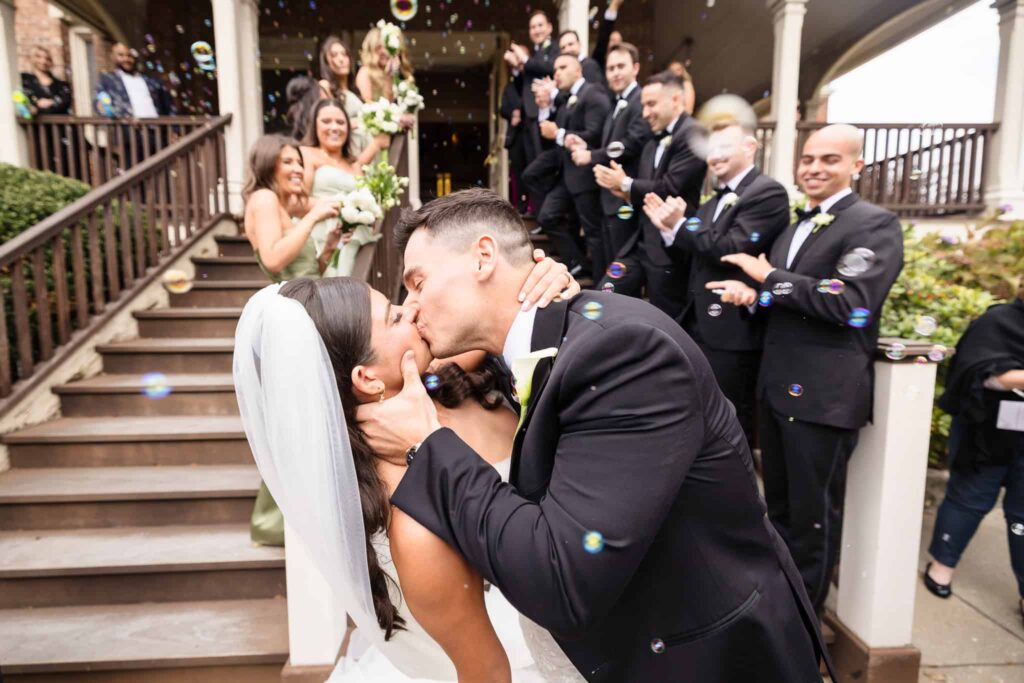 Newlyweds share a joyful kiss surrounded by bubbles and cheers from their bridal party on the church steps, celebrating just after their ceremony at Infant Jesus Church in Port Jefferson.