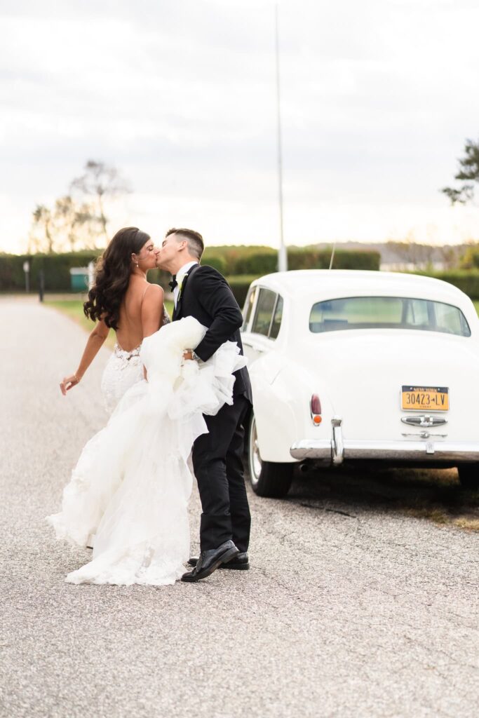 Bride and groom share a kiss beside a vintage white car, her gown flowing in the breeze as they pause for a romantic moment near Old Field Point Lighthouse on Long Island.