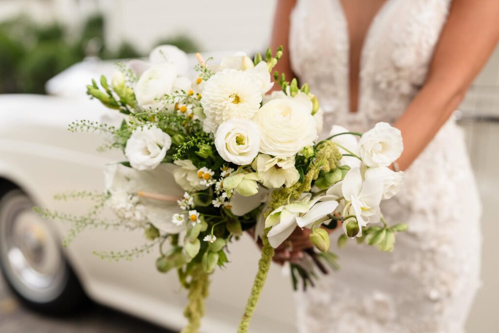 Close-up of the bride’s lush white bouquet featuring orchids, ranunculus, chamomile, and greenery, held in front of her lace gown with a vintage car softly blurred in the background.