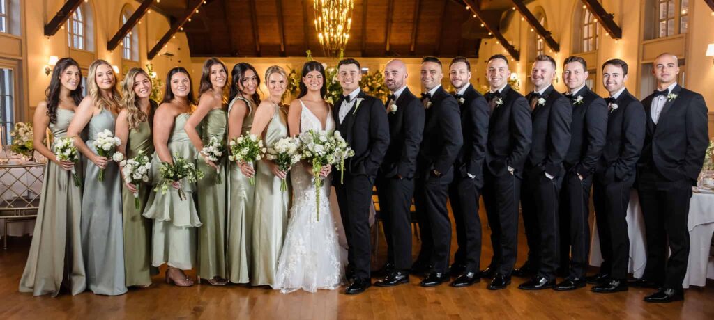 Full wedding party portrait inside the Old Field Club reception hall, with bridesmaids in sage green dresses and groomsmen in black tuxedos, standing with the bride and groom beneath glowing chandeliers and string lights.