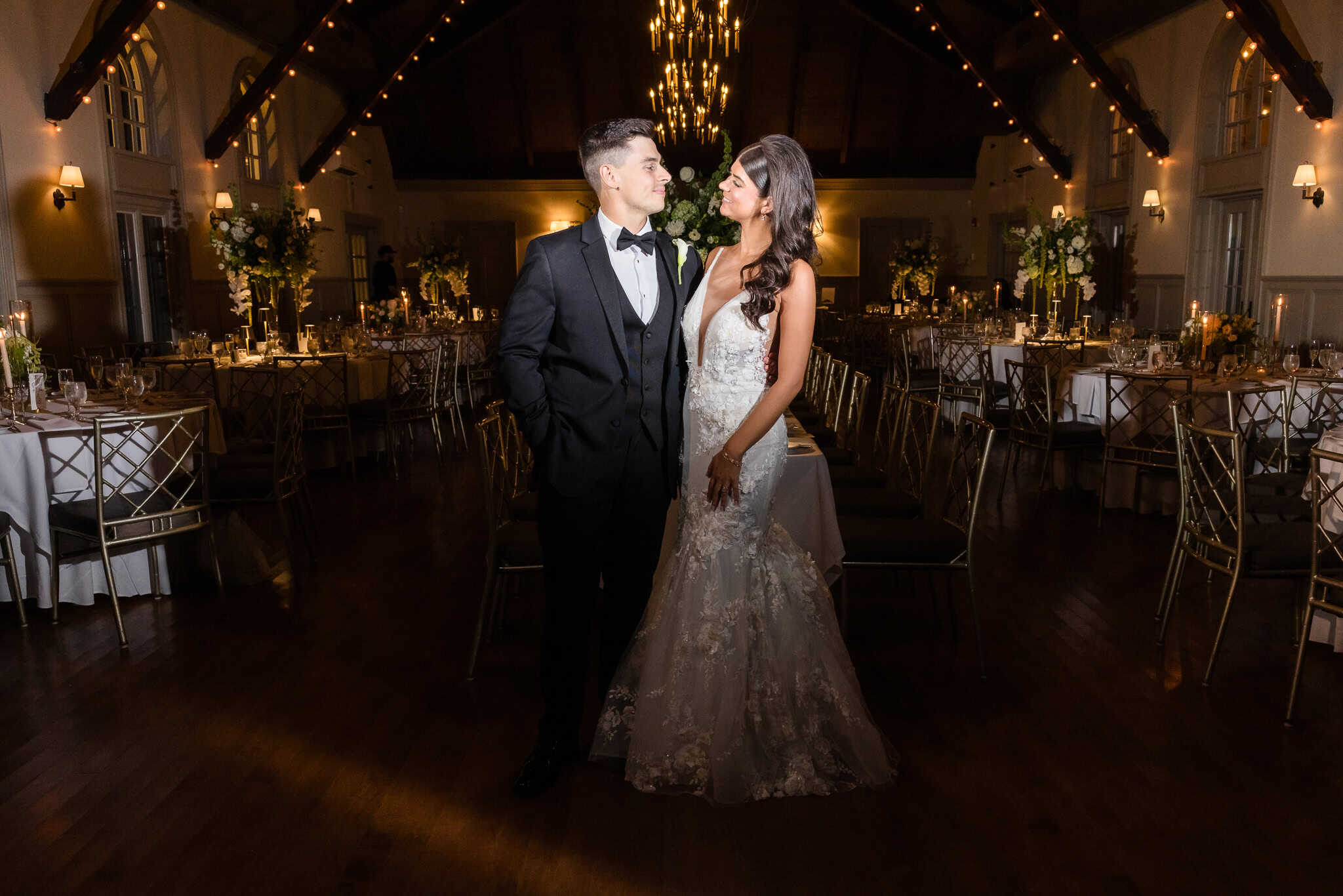 Bride and groom share a quiet, romantic moment in the candlelit Old Field Club reception hall, surrounded by elegant tablescapes, floral arrangements, and warm string lights under a vaulted ceiling.