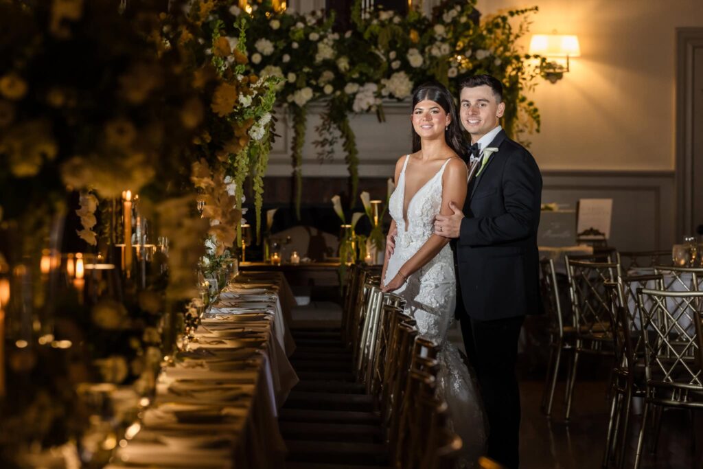 Bride and groom posing beside their elegantly styled king’s table at the Old Field Club, surrounded by cascading florals, candlelight, and refined décor for their intimate Long Island wedding reception.