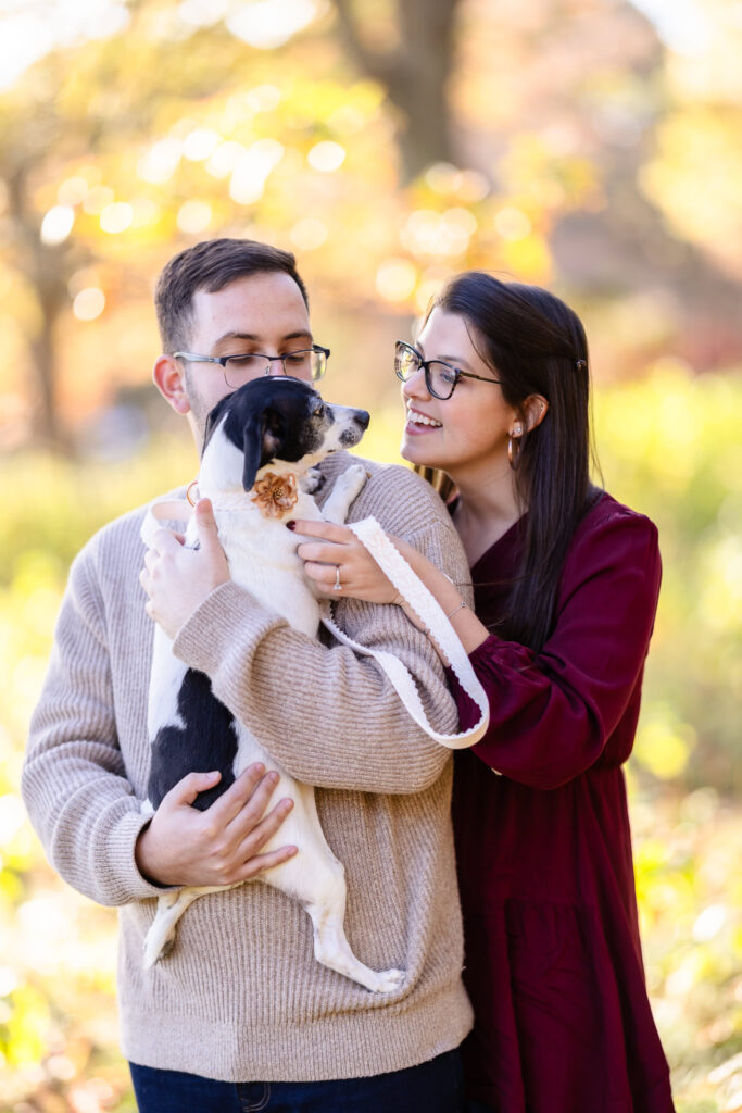 Engaged couple smiling and holding their small dog dressed with a flower collar during a fall engagement session.