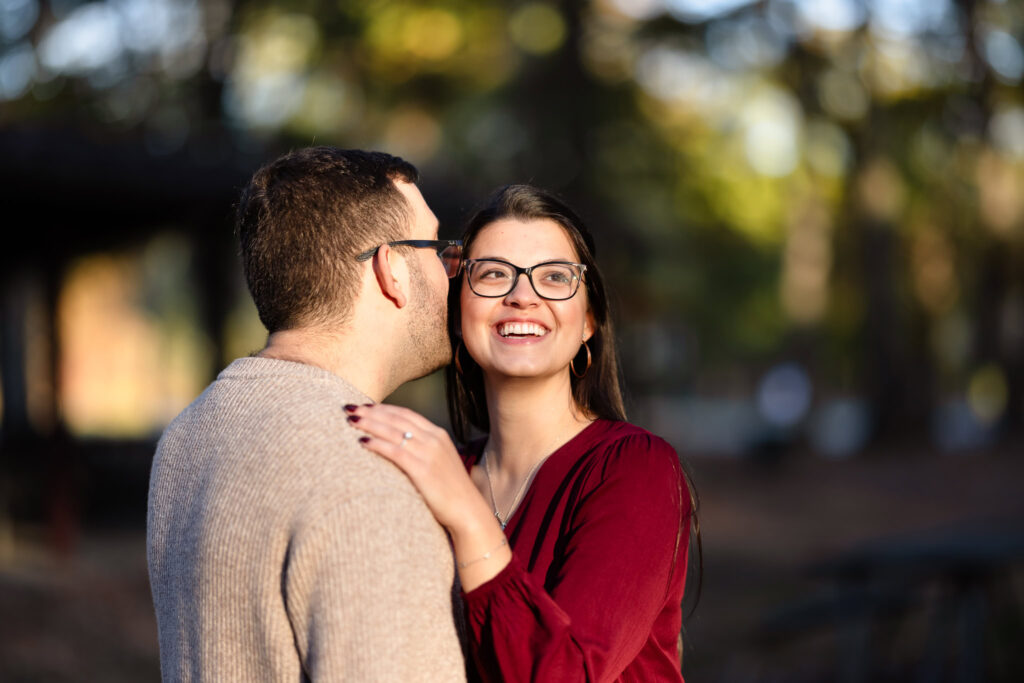 Groom-to-be whispering into his fiancée’s ear as she smiles brightly, captured during a golden hour engagement session.