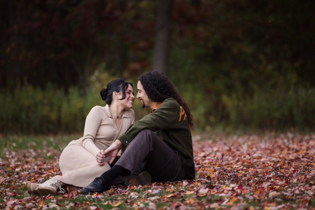 Couple sitting close together on a blanket of colorful fallen leaves at Bethpage State Park, sharing a playful and intimate moment during their fall engagement session.