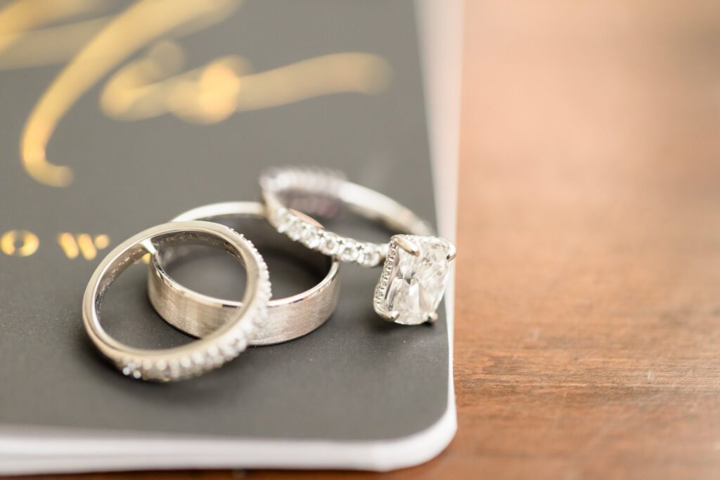 Close-up of wedding rings, including a radiant-cut diamond engagement ring and two white gold bands, styled on vow book for Tellers Next Door wedding detail.