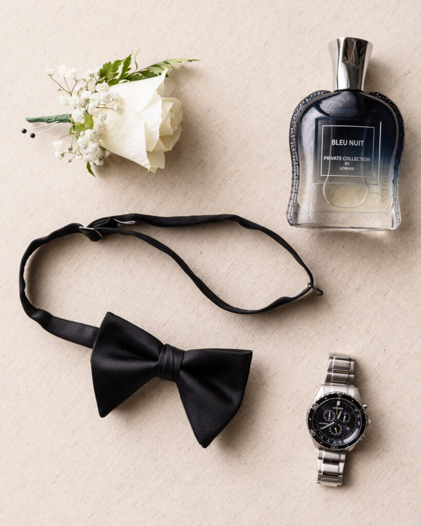 Flat lay of groom’s wedding day details including boutonnière, Bleu Nuit cologne, black bow tie, and stainless steel wristwatch for elegant Tellers Next Door wedding.