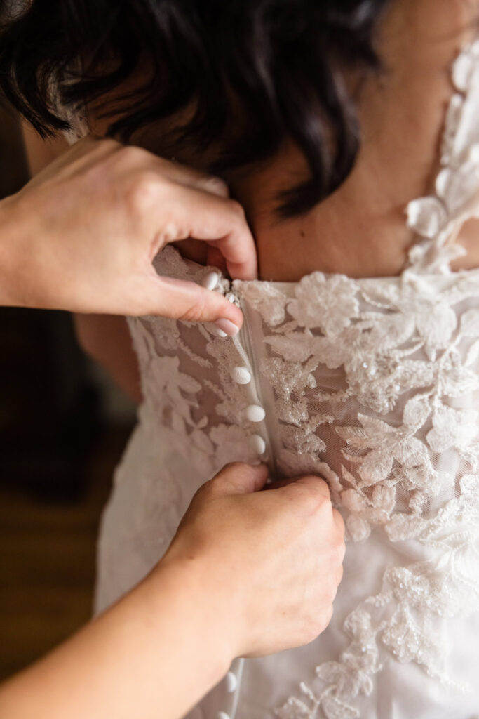 Close-up of hands buttoning intricate lace wedding gown as bride gets ready for elegant Tellers Next Door wedding ceremony.