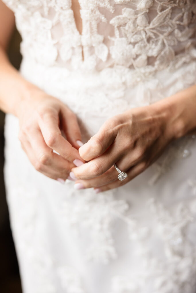Close-up of bride’s hands showcasing her engagement ring and delicate lace details on her wedding dress before the ceremony.