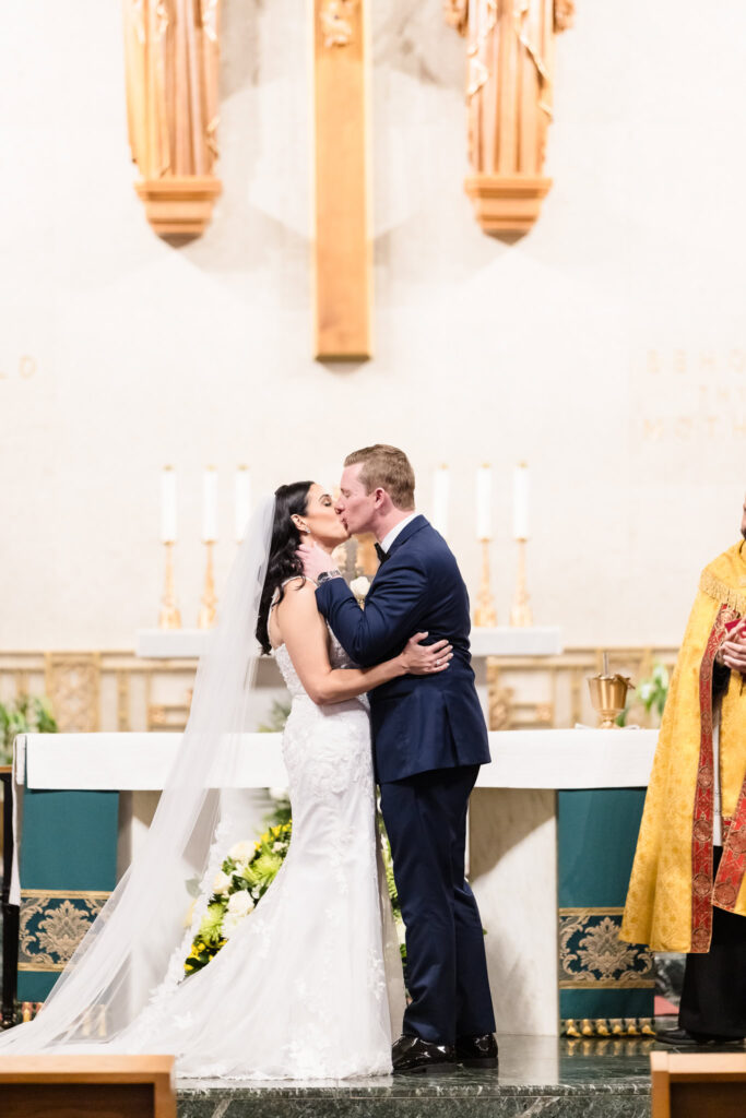 Bride and groom share their first kiss at the altar of Holy Name of Mary Church during emotional Long Island wedding ceremony.