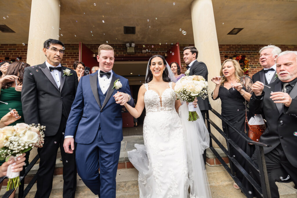 Joyful bride and groom exit the church hand in hand as guests blow bubbles in celebration after their wedding ceremony.