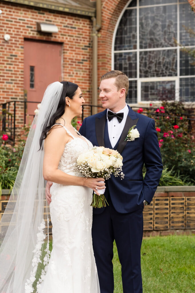 Bride and groom smile at each other outside Holy Name of Mary Church, surrounded by brick architecture and roses after their Long Island wedding ceremony