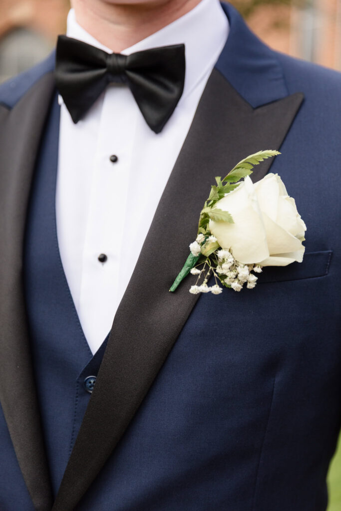 Close-up of groom’s navy tuxedo with black lapels, white rose boutonnière, and classic black bow tie at Tellers Next Door wedding.