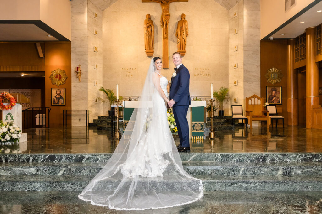Bride and groom hold hands at the altar of Holy Name of Mary Church, with bride’s long lace train flowing behind her in a classic Long Island wedding moment.