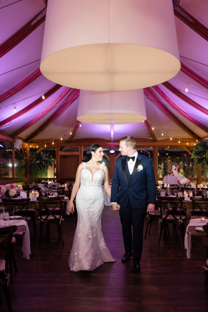 Bride and groom walk hand in hand through their elegantly decorated Tellers Next Door reception space with soft purple uplighting.