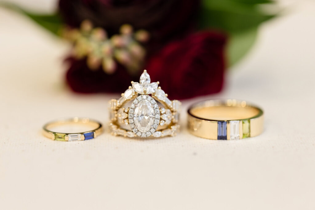 Close-up of ornate marquise-cut diamond engagement ring with coordinating gold wedding bands featuring gemstone inlays, styled against deep red florals.