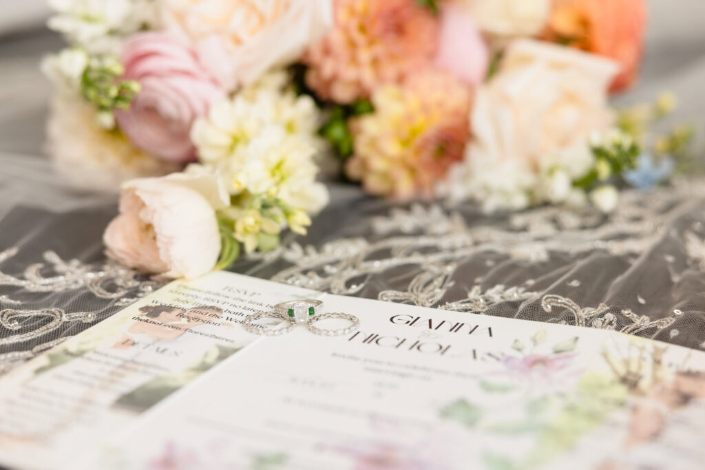 Elegant flat lay of wedding invitation suite with emerald-accented rings, soft floral bouquet, and embroidered bridal veil, styled for a wedding on Long Island.