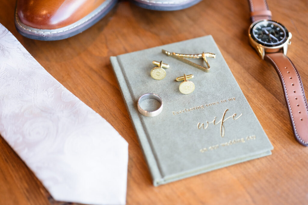 Flat lay of groom’s details including vow book, gold cufflinks, tie clip, watch, and wedding ring, styled with brown leather shoes and white paisley tie.