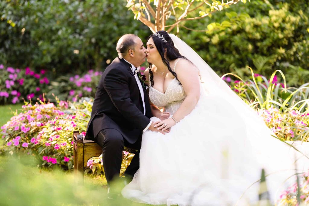 Bride and groom share a kiss while seated on a garden bench surrounded by blooming flowers at Westbury Manor, capturing a romantic portrait on the venue’s manicured estate grounds.