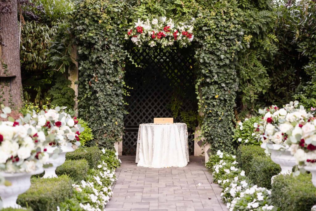Ceremony altar framed by ivy-covered archway and lush florals at Westbury Manor, featuring white and burgundy arrangements lining the aisle for a timeless garden wedding on Long Island.