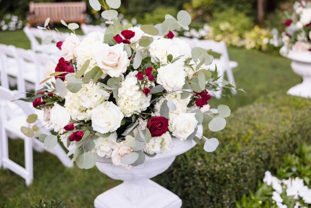 Close-up of a classic garden ceremony floral arrangement with white hydrangeas, ivory roses, and burgundy blooms, styled in a white urn at Westbury Manor on Long Island.