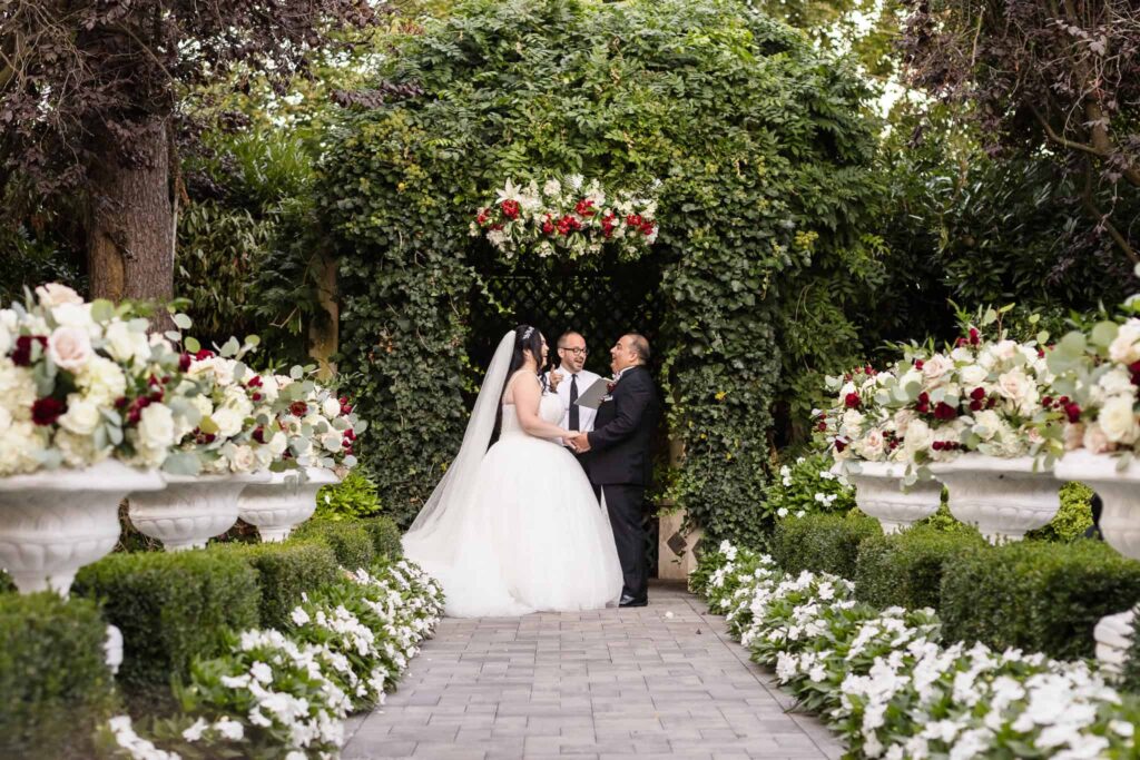 Bride and groom exchange vows beneath a lush ivy-covered archway, surrounded by floral urns and white blooms during a garden ceremony at Westbury Manor on Long Island.