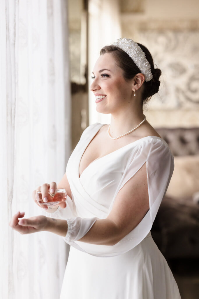 Bride in a classic white gown smiling while applying perfume near a window in the bridal suite at Giorgio’s Baiting Hollow.