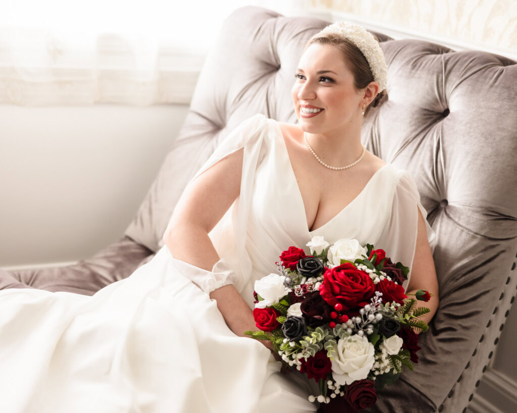 Bride relaxing on a velvet tufted chaise with a winter wedding bouquet of red, white, and black roses.